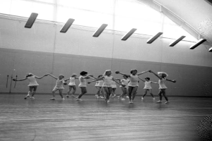 Eine Gruppe junger Mädchen in einem Tanzstudio, die sich an den Händen halten und auf dem Boden tanzen, mit einem Ball auf der rechten Seite, Lampen an der Decke und einem Objekt an der Wand im Hintergrund, in Schwarz-Weiß.