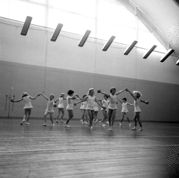 Eine Gruppe junger Mädchen in einem Tanzstudio, die sich an den Händen halten und auf dem Boden tanzen, mit einem Ball auf der rechten Seite, Lampen an der Decke und einem Objekt an der Wand im Hintergrund, in Schwarz-Weiß.
