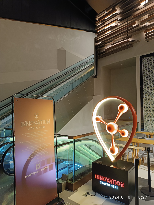 Großes Schild mit der Aufschrift "Innovation beginnt hier" in einem Gebäude mit einer Rolltreppe im Hintergrund, Tischen und Stühlen auf der rechten Seite und einer Tafel auf der linken Seite, die die Teilnahme an der HSTP20 Expo anzeigt.