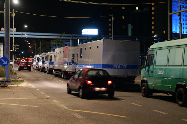 Eine lange Reihe von Polizeiwagen, die an einer Straße bei Nacht geparkt sind, mit Verkehr, Straßenschildern, Versorgungsmasten, Gebäuden und einer Brücke im Hintergrund.