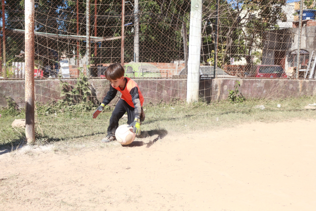 Junger Junge in Fußballschuhen beim Spielen auf einem Schmutzfeld mit Gras, Pflanzen, Pfosten, einem Zaun, einer Wand, Bäumen, Fahrzeugen, Gebäuden und einem bewölkten Himmel im Hintergrund.