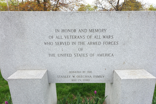 Gedenkstein mit einer Inschrift für US-Streitkräfte-Veteranen, umgeben von Pflanzen, Blumen, Gras, Bäumen und einem bewölkten Himmel.