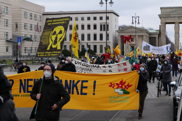 Große Gruppe von Menschen marschiert auf einer Straßendemonstration gegen Atomkraft in Deutschland, tragend Schilder und Fahnen, mit Fahrzeugen auf der rechten Seite und Gebäuden, Laternenpfählen, Bäumen und einem Tor im Hintergrund.