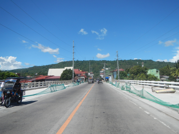 Autobahn mit fahrenden Fahrzeugen, ein geparktes Motorrad auf dem Seitenstreifen, Geländer, Strommasten mit Drähten, Bäume, Gebäude, ferne Hügel und ein bewölkter Himmel.