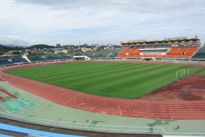 Ein großes Stadion mit einem Fußballfeld, umgeben von Gebäuden, Bäumen, Hügeln und einem klaren blauen Himmel, mit ein paar Menschen und saftigem Grün.