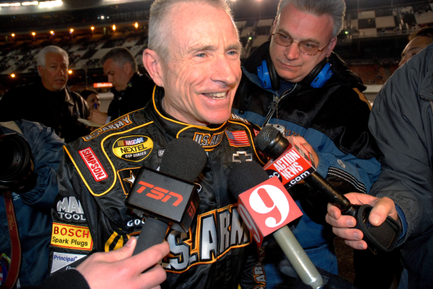 Ein Mann in einer schwarzen Jacke spricht mit Reportern vor einem hell erleuchteten Stadion voller Zuschauer.