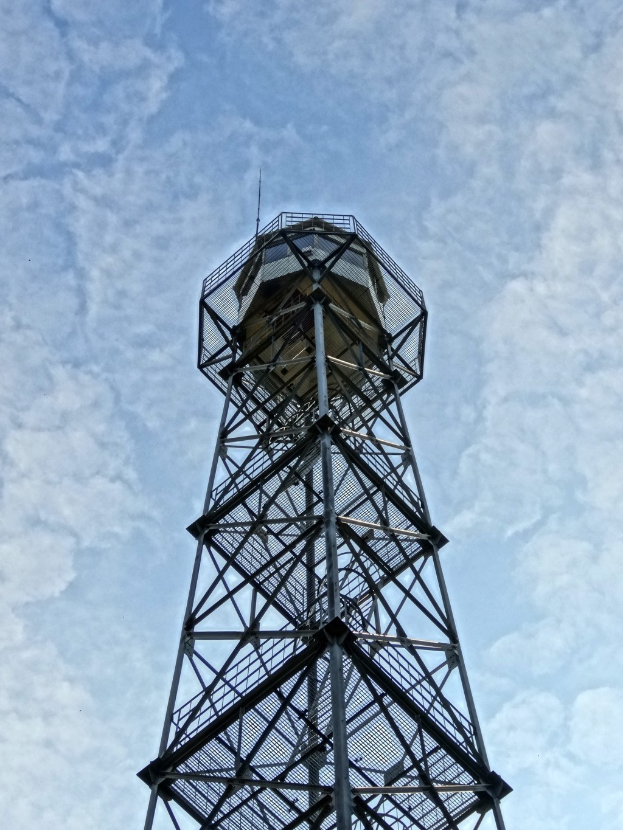 Ein hoher Metallfeuerturm steht vor einem blauen Himmel mit weißen, flauschigen Wolken.