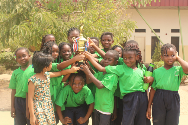 Eine Gruppe von Kindern hält einen Pokal vor einem Schulgebäude mit Bäumen und Pflanzen im Hintergrund