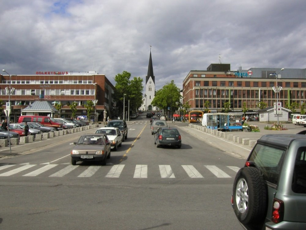 Stadtstraße mit parkenden Autos, Gebäuden, Bäumen, Laternenmasten und einem bewölkten Himmel, mit einem sichtbaren Kfz-Kennzeichen.