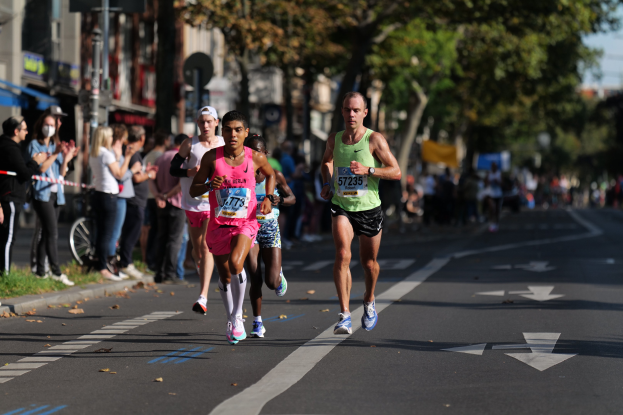 Gruppe von Läufern bei einem Stadtmarathon, mit Zuschauern links, unscharfen städtischen Hintergrund mit Bäumen, Gebäuden und einem Fahrrad.