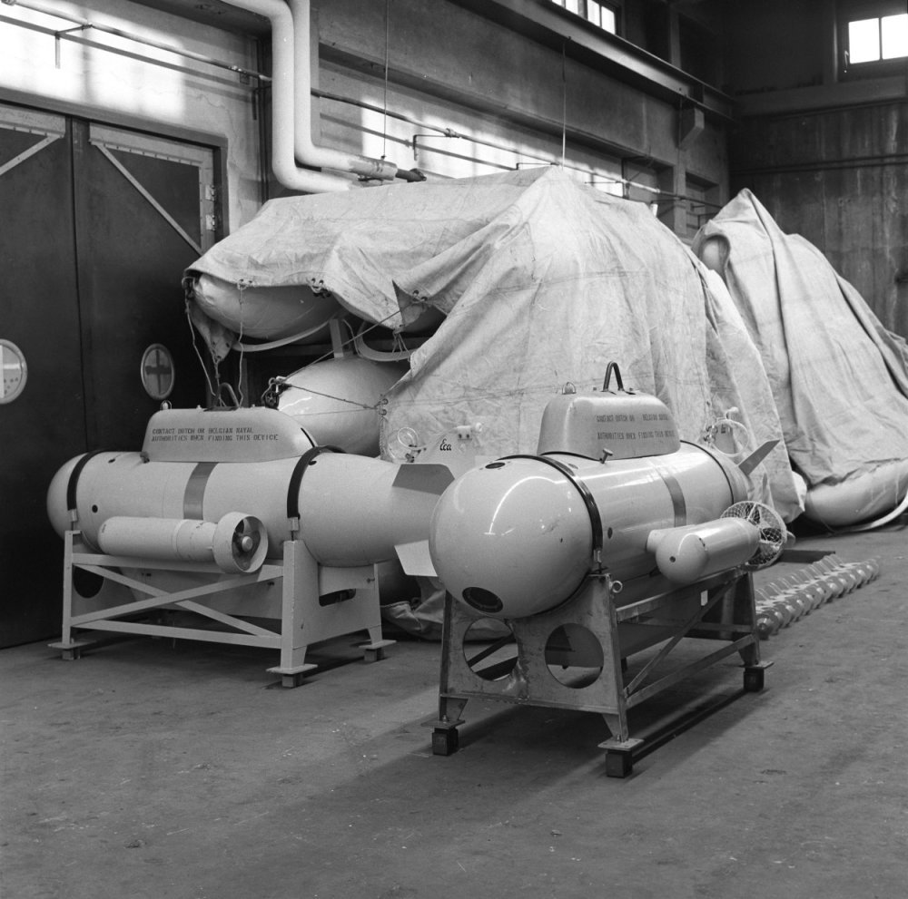 Ein Schwarz-Weiß-Foto eines Raums, der mit verschiedenen Tanktypen gefüllt ist, darunter eine Wasserstoffbombe, mit einer Wand, einer Tür, Fenstern, Rohren und anderen Gegenständen im Hintergrund.