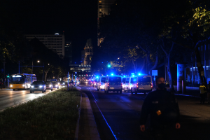 Eine Stadtstraße bei Nacht mit Polizeiwagen, Bussen, Fußgängern auf dem Gehweg, Gras, Bäumen, Gebäuden und Laternenmasten.