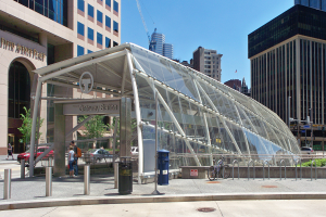 Eine belebte Stadtstraße mit hohen Wolkenkratzern, eine Bushaltestelle mit Glasdach, Menschen, Fahrzeuge, ein Metallzaun, ein Mülleimer, Pfosten, ein Schild, eine Ampel, Bäume, ein Kran und ein bewölkter Himmel.