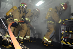 Feuerwehrleute in Schutzausrüstung arbeiten in der Nähe eines Löschfahrzeugs mit einem Gestell von Gegenständen und Hintergrundrohren und -leuchten.
