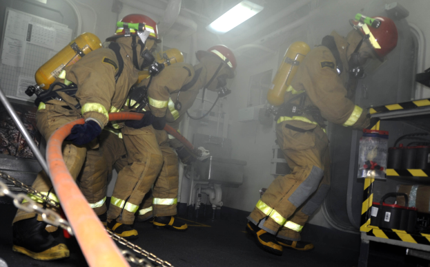 Feuerwehrleute in Schutzausrüstung arbeiten in der Nähe eines Löschfahrzeugs mit einem Gestell von Gegenständen und Hintergrundrohren und -leuchten.