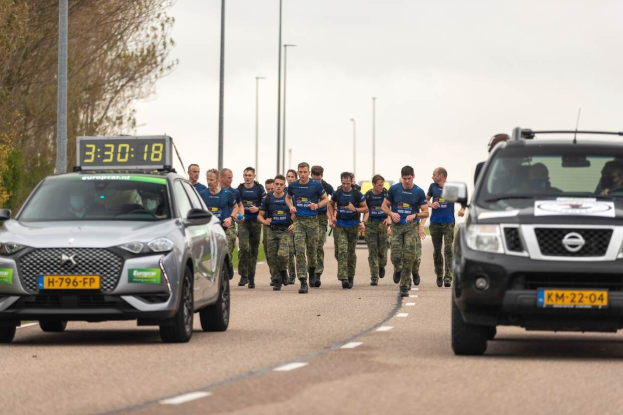 Gruppe von Menschen, die auf einer Straße neben geparkten Autos mit Bäumen auf der linken Seite und Strommasten im Hintergrund laufen, unter einem sichtbaren Himmel, an einer Europäischen Cross-Country-Challenge teilnehmend.