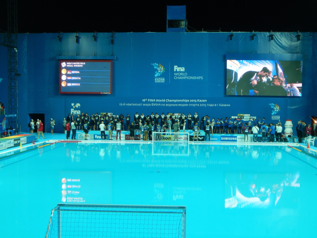 Eine Gruppe von Menschen um ein Hallenbad mit einem Netz am Boden herumstehend, mit einer Wand im Hintergrund, die "FINA World Championships 2015 Kazan"-Text und Logos anzeigt, sowie Gerüste und Beleuchtung.