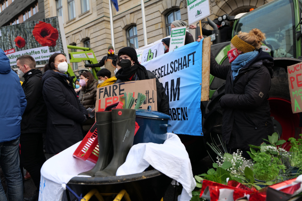 Maskierte Menschen mit Protestschildern vor einem Lastwagen mit einem Tisch, Pflanzen und Gebäuden im Hintergrund.
