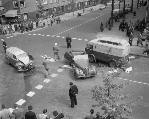 Eine Schwarz-Weiß-Szene eines Autounfalls an einer Straßenkreuzung mit mehreren Fahrzeugen, darunter ein Van, und einer Gruppe von Menschen, die darum herumstehen, mit Gebäuden, Bäumen, Pfählen und Texttafeln im Hintergrund.