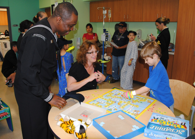 Eine Gruppe von Menschen, die ein Brettspiel an einem Tisch mit Spielzeugen, Boxen und anderen Gegenständen spielen, mit Schränken, Salzflaschen, einer Tür und einer Wand im Hintergrund.