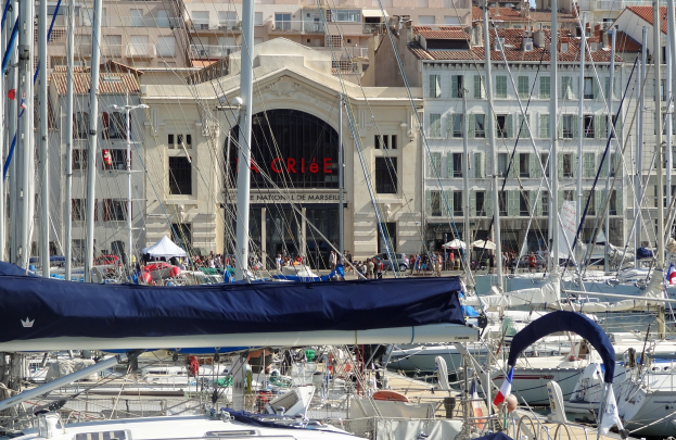 Ein Hafen mit zahlreichen Booten im Wasser, Gebäude mit Fenstern und einem Namensschild im Hintergrund sowie Zelte mit anwesenden Menschen.