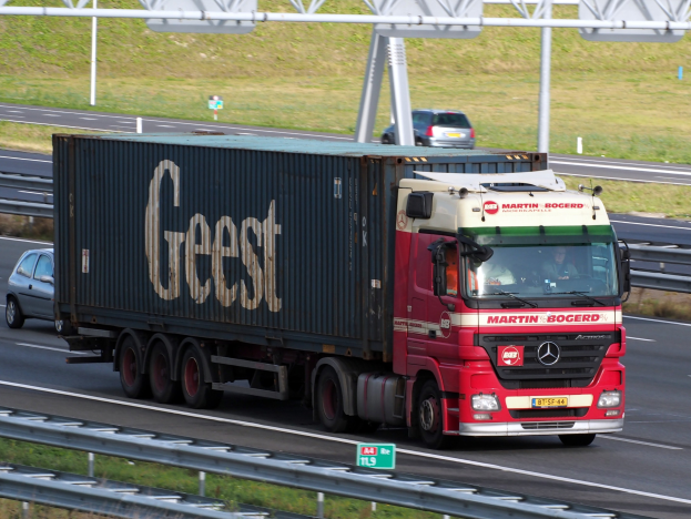 Roter Lastwagen fährt auf einer Autobahn mit einem Container auf dem Rücken, der ein paar Menschen trägt, mit Strommasten, Holzplanken und Gras im Hintergrund.