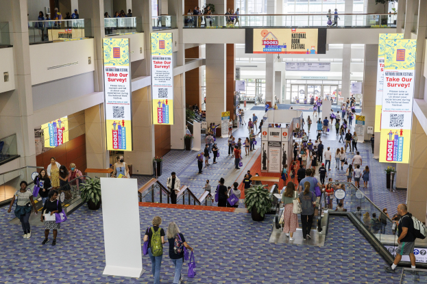 Eine große Gruppe von Menschen geht durch ein großes Gebäude mit Transparenten, Topfpflanzen, Rolltreppen, Geländern, Säulen, bedruckten Schildern, Glastüren und anderen Gegenständen, einige tragen Taschen, wahrscheinlich nehmen sie am Nationalen Buchfest teil.