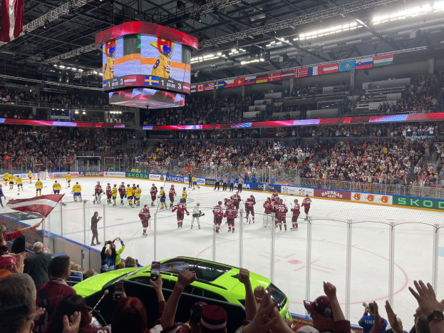 Ein Eishockeyspiel in einer großen Arena mit Zuschauern, einige halten Fahnen, ein Auto in der Nähe, Bande, Banner, Deckenbeleuchtung, Bildschirm und hängende Fahnen.