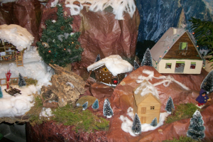Ein Miniaturdorf-Modell mit einem zentralen Weihnachtsbaum, umgeben von schneebedeckten Häusern, Bäumen, Pflanzen, Felsen und anderen Gegenständen, mit einer Wand im Hintergrund.