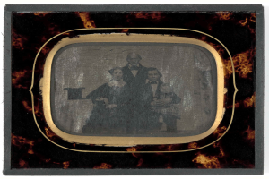 Ein altes Foto eines Mannes im Anzug und einer Frau in unbekannter Kleidung, die vor einem Fernseher stehen und ernst schauen.