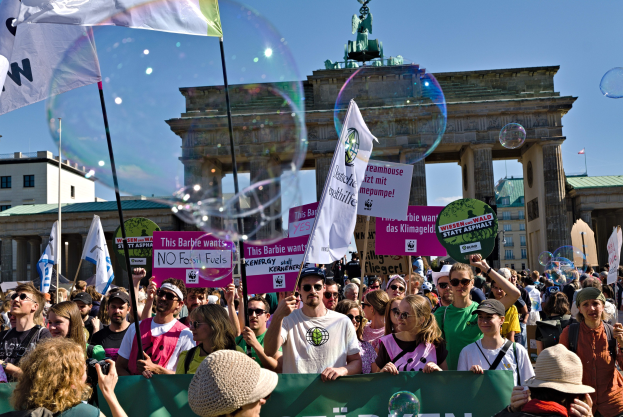 Menschenmenge vor dem Brandenburger Tor in Berlin, viele mit Mützen und Schutzbrillen, einige mit Fahnen und Schildern mit Text, im Hintergrund Gebäude, ein Tor mit einer Statue darauf und ein Himmel voller Seifenblasen.