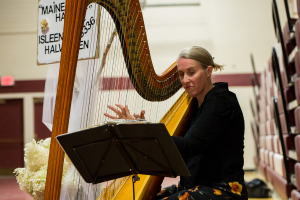 Eine Frau sitzt da und schaut in ein Buch vor ihr, während sie eine Harfe spielt. Links von ihr ist eine Tafel, und im Hintergrund gibt es Türen mit einem Ausgangsschild. Unten steht ein Blumenstrauß neben einem Vorhang, und rechts ist eine Wand.