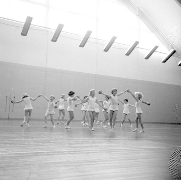 Schwarzes und weißes Bild von jungen Mädchen, die sich an den Händen halten und in einem Studio tanzen, mit einem Ball auf der rechten Seite und Lampen an der Decke, mit einem Objekt an der Wand im Hintergrund.