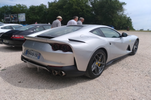 Ein roter Ferrari 812 Superfast wird auf dem Goodwood Festival of Speed gezeigt, umgeben von Zuschauern mit Mützen, Bäumen, Informationsschildern und einem bewölkten Himmel im Hintergrund.