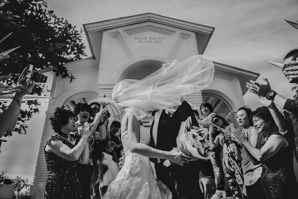 Ein Brautpaar steht in der Mitte, mit einer Person, die links eine Kamera hält, einem Gebäude im Hintergrund, einem Baum und einem Strauß im Bild, unter einem Himmel.