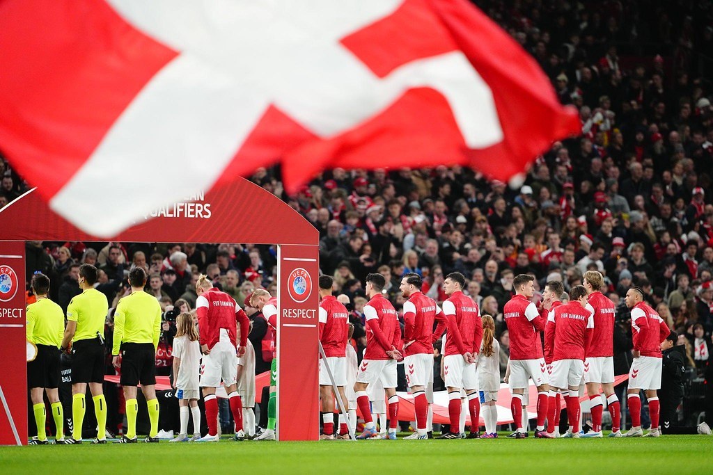 Gruppe von Menschen auf einem Fußballfeld mit einer roten und weißen Flagge im Vordergrund, einem Bogen mit 'Bayern Munich vs Bayern Munich Betting Tips & Preview' im Hintergrund und einer großen Menge im Stadion.