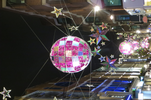 Eine Stadtstraße bei Nacht mit zahlreichen Lichtern und Dekorationen, darunter eine große Diskokugel, die von einem Gebäude hängt und eine festliche Stimmung schafft.