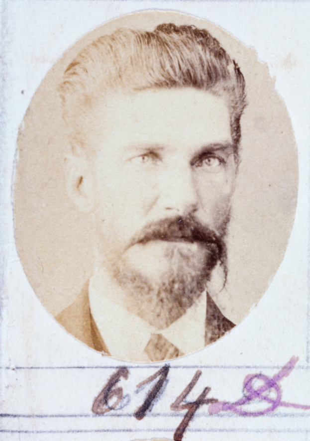 Ein altes Foto eines Mannes mit Bart und Schnurrbart, der direkt in die Kamera schaut und dessen Lippen leicht geöffnet sind.