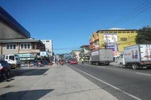 Eine belebte Stadtstraße mit zahlreichen Autos, Lastwagen, Fußgängern, Gebäuden, Schildern, Strommasten mit Drähten, Bäumen und einem bewölkten Himmel.