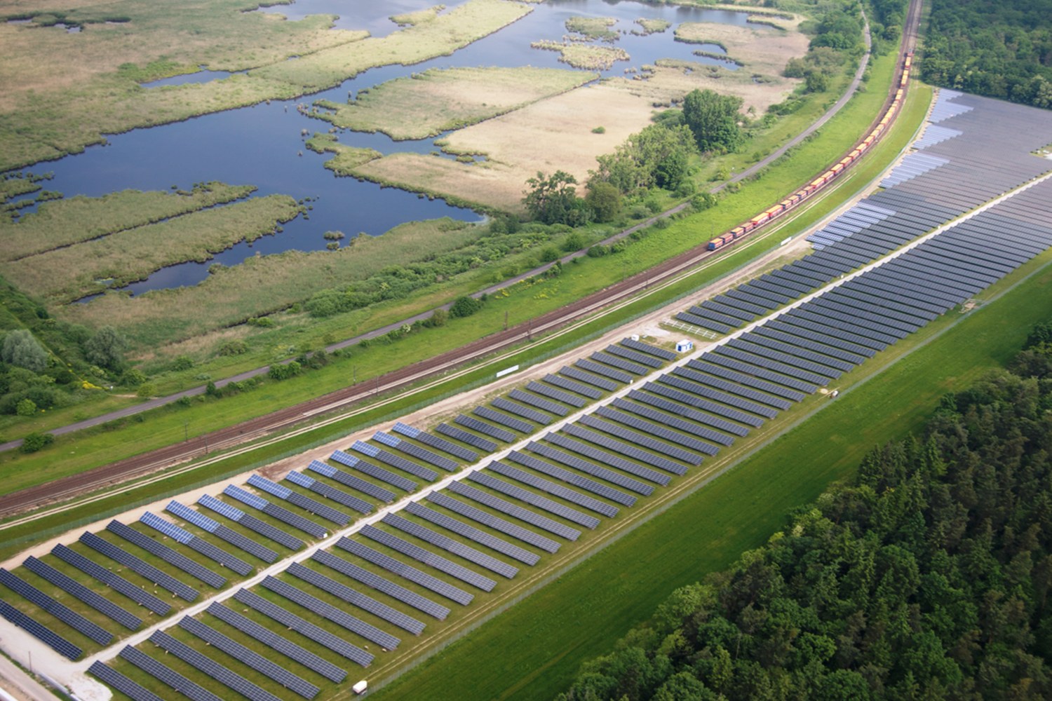 Luftaufnahme einer Solaranlage mit Solarpanelen in einem Feld, umgeben von Bäumen, Gras, Pflanzen und Wasser, mit einem Zug auf einer nahen Bahnlinie.