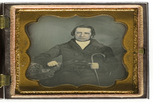 Ein altes Daguerreotyp eines Mannes in einem Anzug, der in einem Stuhl sitzt und einen Stock hält, mit ernstem Gesichtsausdruck und konzentriertem Blick.