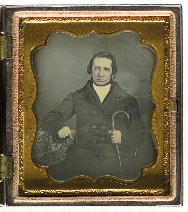 Ein altes Daguerreotyp eines Mannes in einem Anzug, der in einem Stuhl sitzt und einen Stock hält, mit ernstem Gesichtsausdruck und konzentriertem Blick.