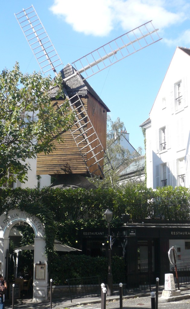 Eine Windmühle, mehrere Gebäude, Bäume, Metallstangen und ein Zaun.