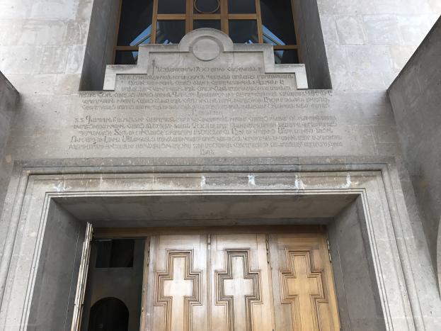 Große hölzerne Tür vor einem steinernen Gebäude mit einem Glasfenster darüber; Inschrift auf der Wand, die wahrscheinlich Holocaust-Opfern gewidmet ist.