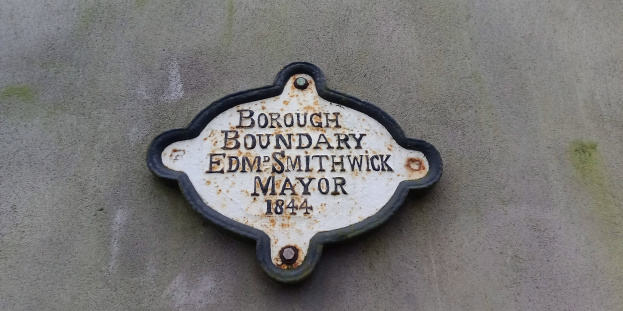 Metallplatte an der Seite eines Gebäudes mit schwarzem Hintergrund und weißer fettgedruckter Schrift, die "Gemeindegrenze Edm Smithwick Bürgermeister" lautet.