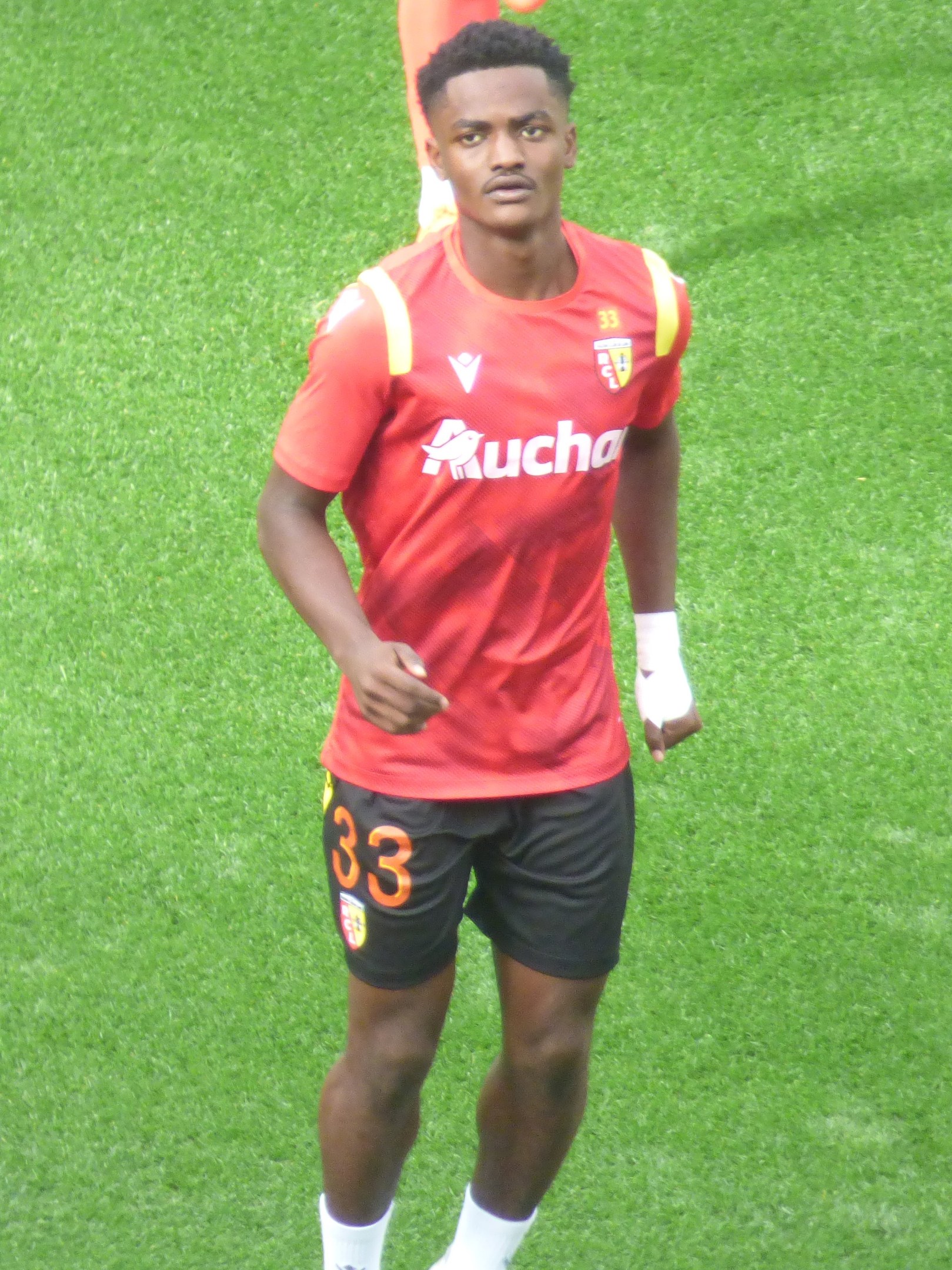 Emmanuel Adebayor in einem roten Trikot und schwarzen Shorts auf einem Fußballfeld mit zwei Personen im Hintergrund.