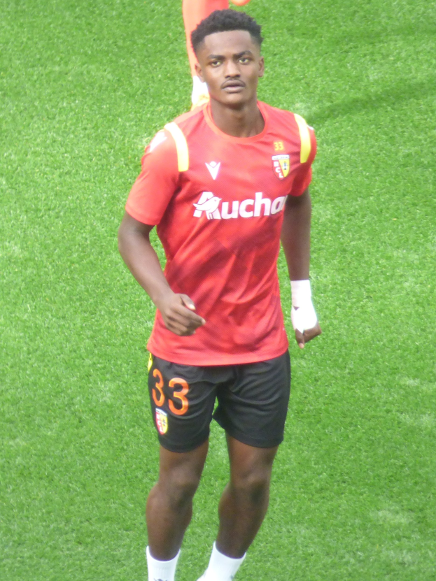 Emmanuel Adebayor in einem roten Trikot und schwarzen Shorts auf einem Fußballfeld mit zwei Personen im Hintergrund.