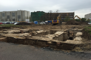 Großer Erdberg an einer Baustelle umgeben von Baggern, Fahrzeugen, Gras, Bäumen und Gebäuden mit Fenstern in einem bewölkten Himmel über einer Straße.