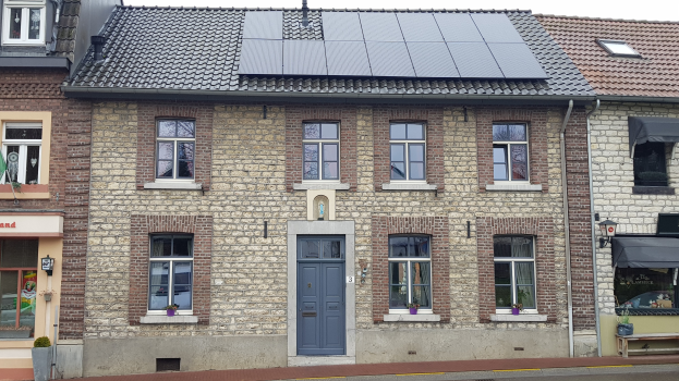 Ein Haus mit Solarpanelen auf dem Dach, das Fenster, Türen, Topfpflanzen, eine Bank, ein Schild und einen Straßenpfosten zeigt, unter einem bewölkten Himmel.