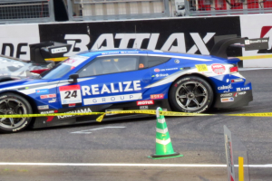 Ein blau-weißer Rennwagen fährt auf einer Rennstrecke, mit einem Verkehrskegel im Vordergrund und Tafeln mit Text, Geländern und anderen Gegenständen im Hintergrund.